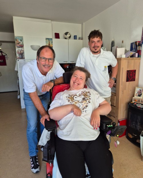 Leonie sitzt im Rollstuhl und hinter ihr stehen Jürgen Menges und Augustin Mare von Handi-Move. Alle lachen in die Kamera.