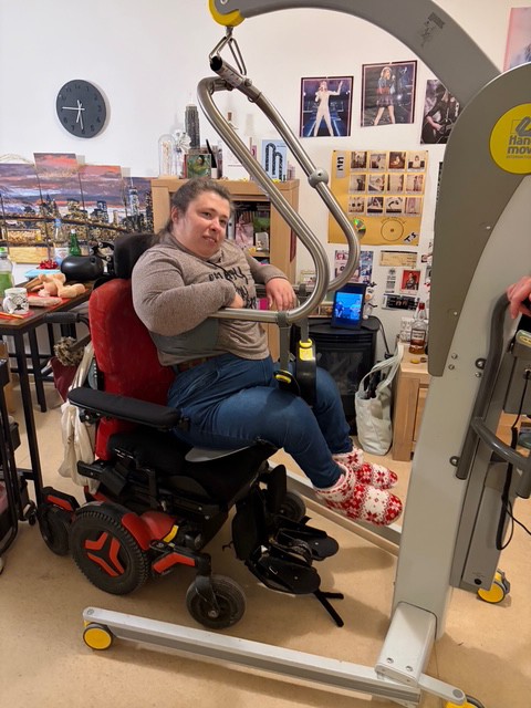 Leonie sitzt im Hebebügel und schwebt über dem Rollstuhl. Der Hebebügel hängt am mobilen Lifter.