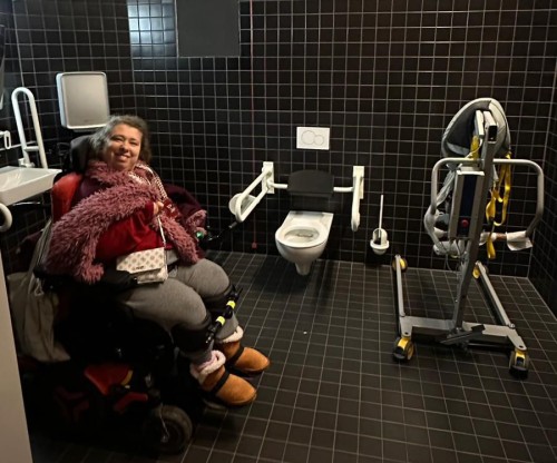schwarz gekachelter Toilettenraum, Leonie sitzt links im Rollstuhl und rechts steht der Reiselifter neben dem WC.